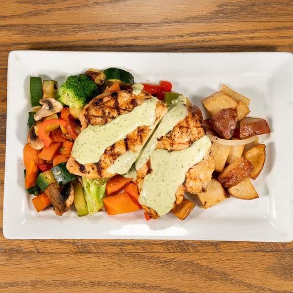 Grilled chicken breasts with green sauce, roasted vegetables, and potatoes on a white rectangular plate.