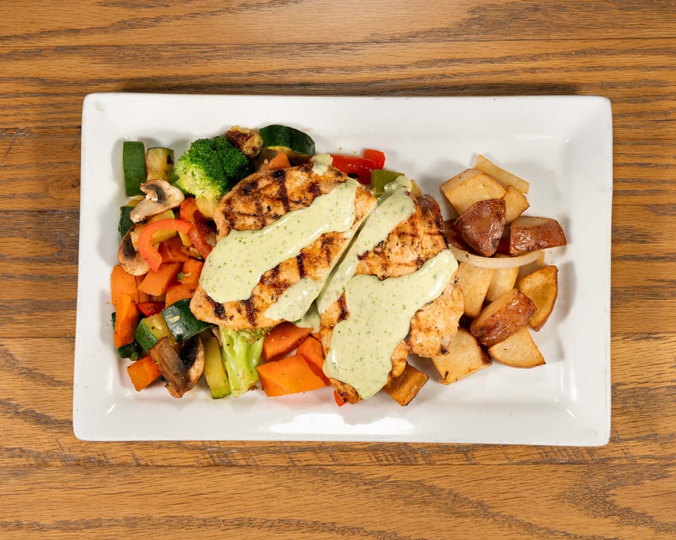Grilled chicken breasts with green sauce, roasted vegetables, and potatoes on a white rectangular plate.