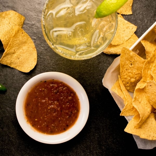 Overhead view of tortilla chips, salsa, jalapeños, and a margarita on a dark surface.