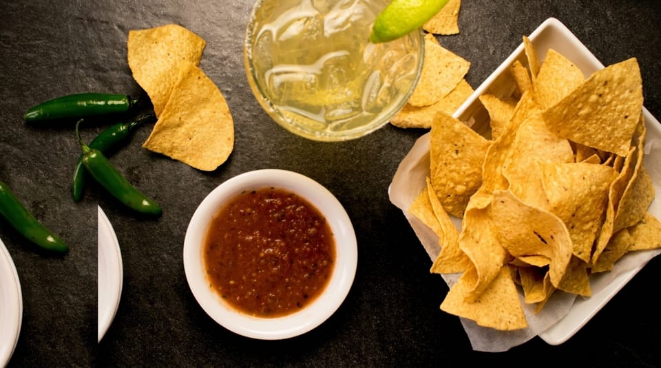 Overhead view of tortilla chips, salsa, jalapeños, and a margarita on a dark surface.