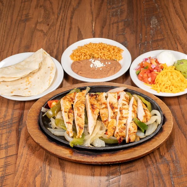 Chicken fajitas sizzling on a cast iron skillet with sides of rice, beans, tortillas, and toppings.