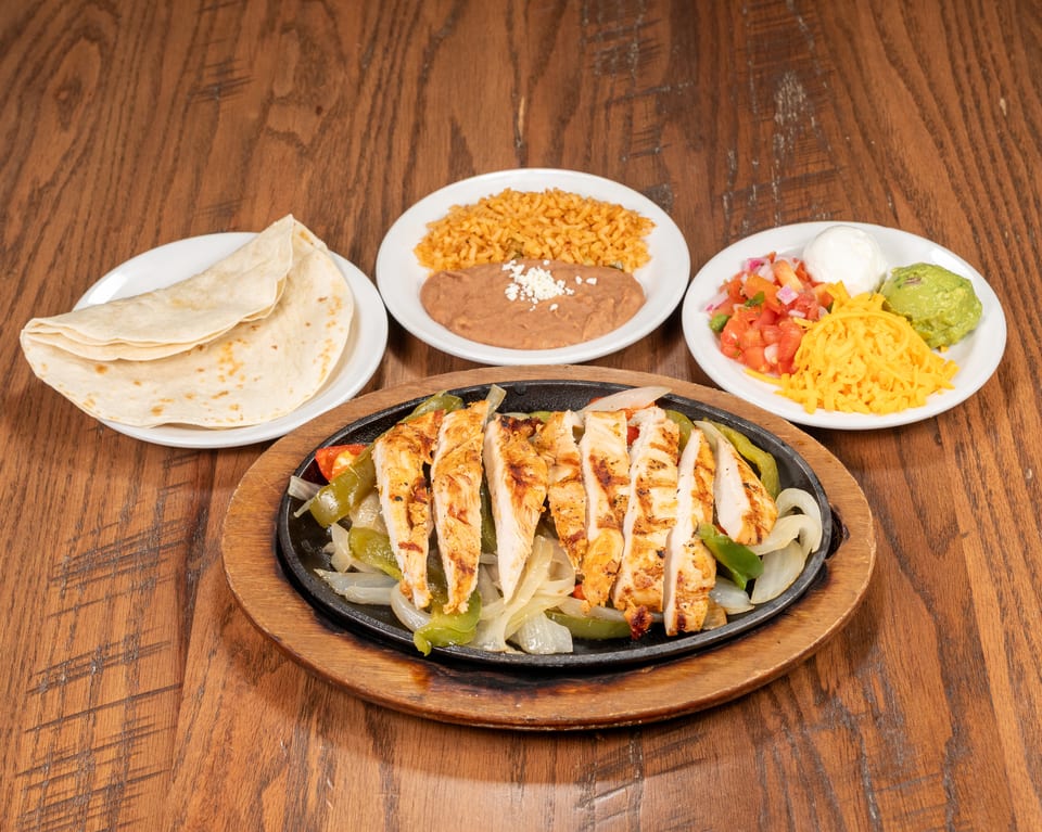 Chicken fajitas sizzling on a cast iron skillet with sides of rice, beans, tortillas, and toppings.