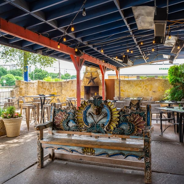 Outdoor patio with a colorful carved wooden bench, tables, chairs, and a stone fireplace under a covered roof.