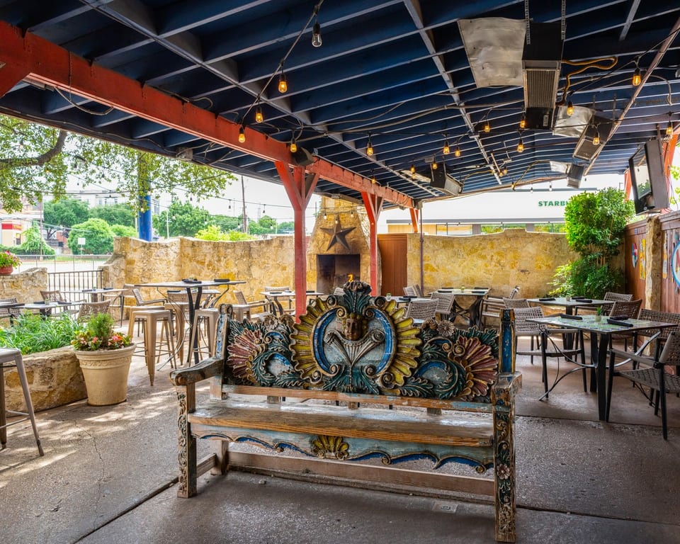 Outdoor patio with a colorful carved wooden bench, tables, chairs, and a stone fireplace under a covered roof.
