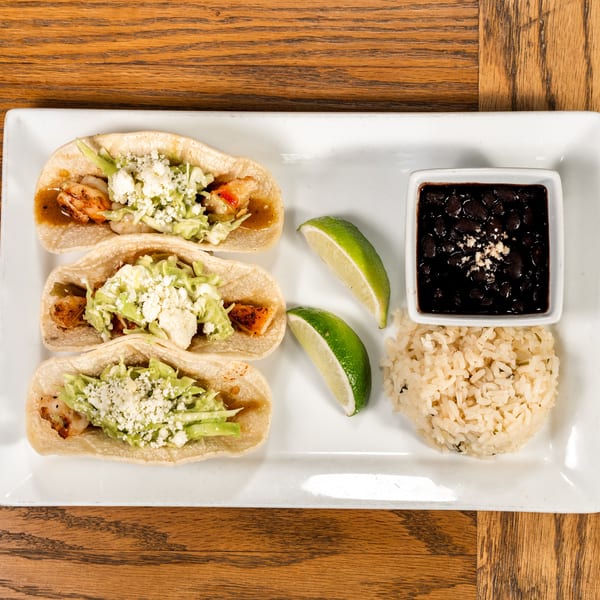 Three shrimp tacos with cabbage, cheese, and sauce, served with rice, black beans, and lime wedges.