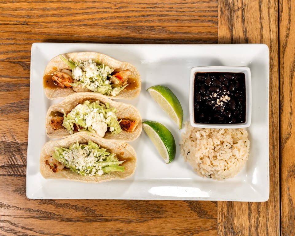 Three shrimp tacos with cabbage, cheese, and sauce, served with rice, black beans, and lime wedges.