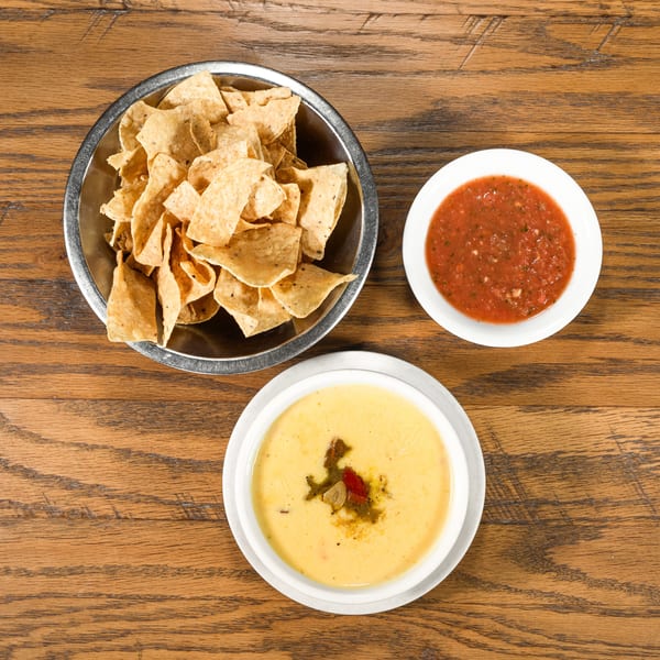 Overhead view of a metal bowl of tortilla chips, a white bowl of salsa, and a white bowl of queso on a wooden table.