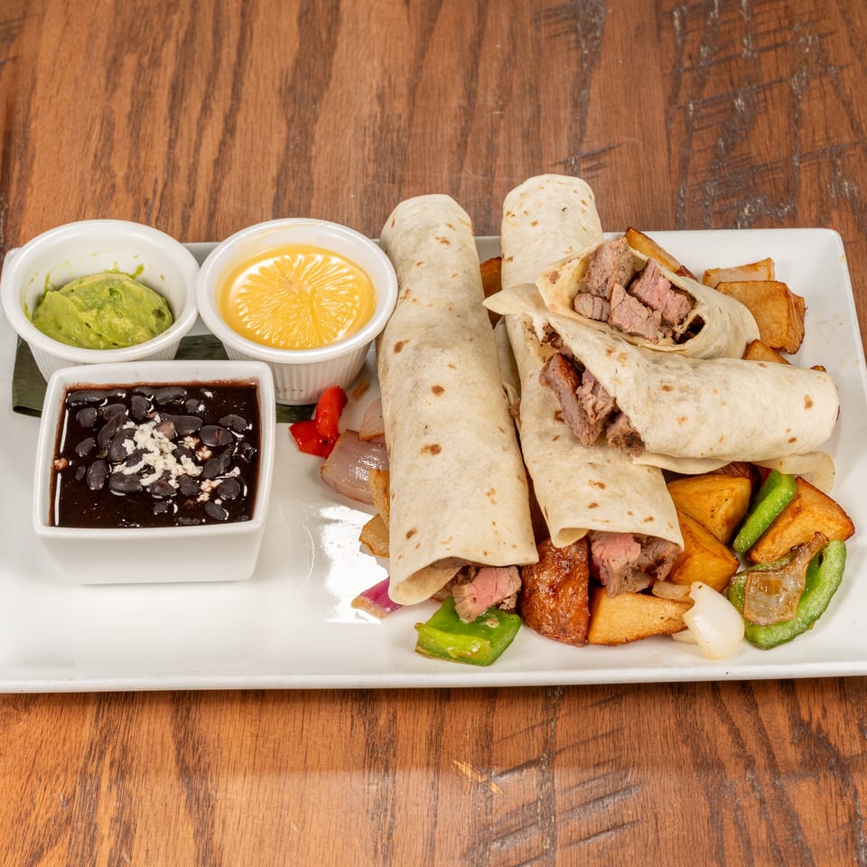 Steak fajita wraps with roasted vegetables, black beans, guacamole, and cheese sauce on a white plate.