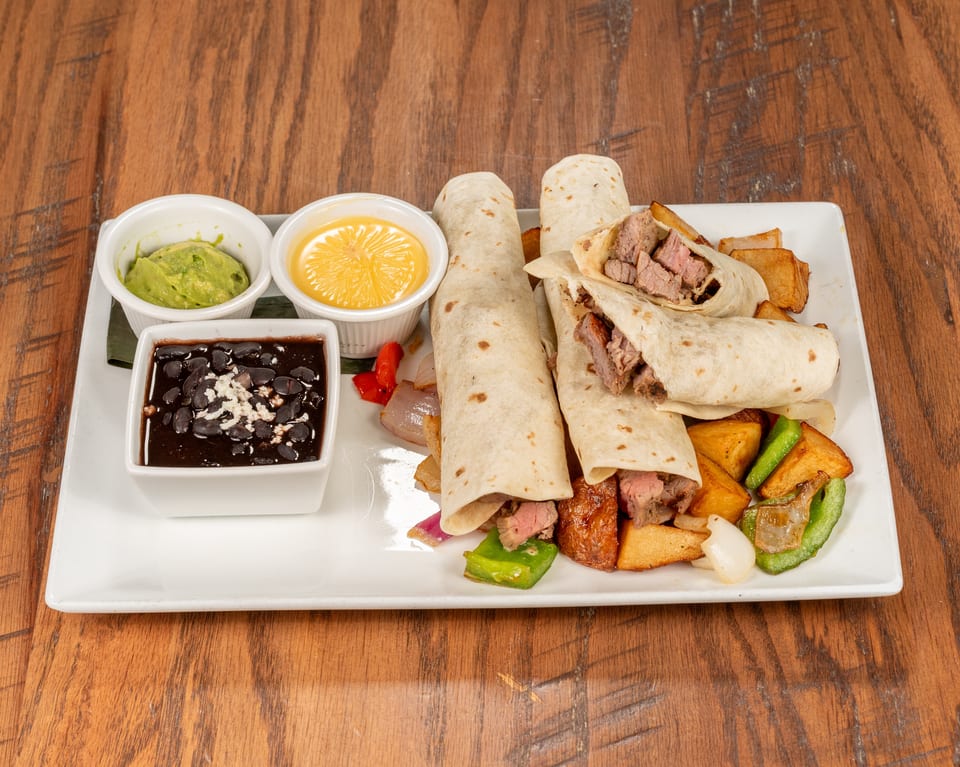 Steak fajita wraps with roasted vegetables, black beans, guacamole, and cheese sauce on a white plate.