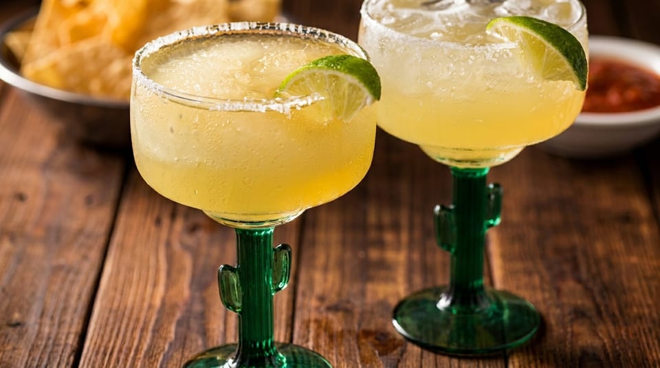 Two frosty margaritas in cactus-stemmed glasses with lime wedges, salt rims, and tortilla chips.