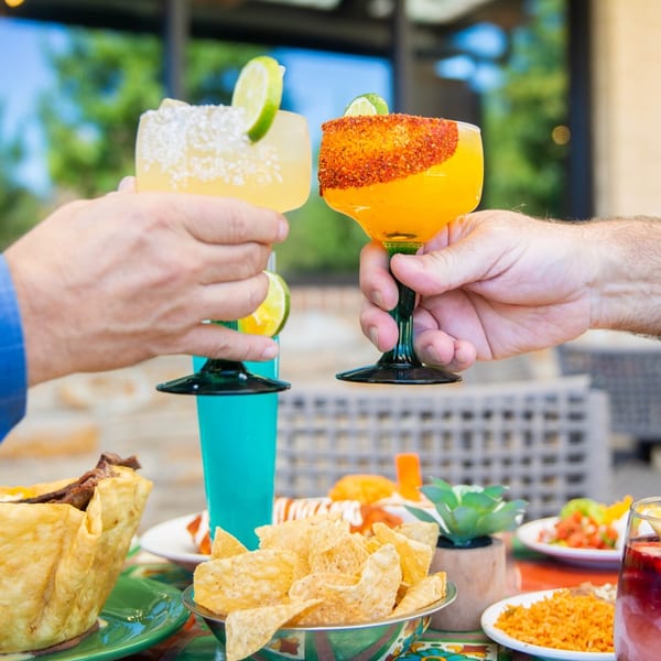 Two people toasting margaritas, one with salt and lime, the other with chili powder, over a table of Mexican food.