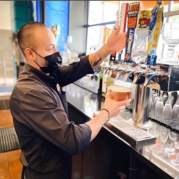 A masked bartender in a black shirt pours a beer from a tap at a bar.