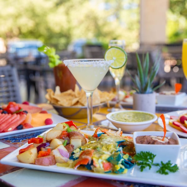 A vibrant outdoor brunch spread with margaritas, mimosas, fruit plates, and Mexican entrees on a colorful tiled table.