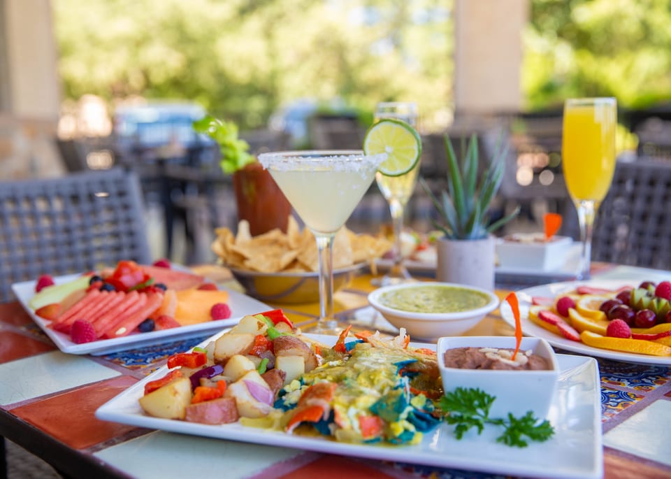 A vibrant outdoor brunch spread with margaritas, mimosas, fruit plates, and Mexican entrees on a colorful tiled table.