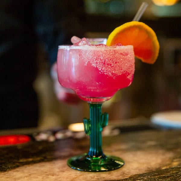 A vibrant pink margarita in a cactus-stemmed glass, garnished with an orange slice and straw.