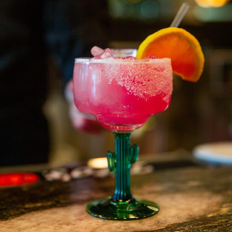 A vibrant pink margarita in a cactus-stemmed glass, garnished with an orange slice and straw.
