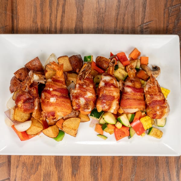 Bacon-wrapped shrimp with roasted potatoes and mixed vegetables on a white rectangular plate.