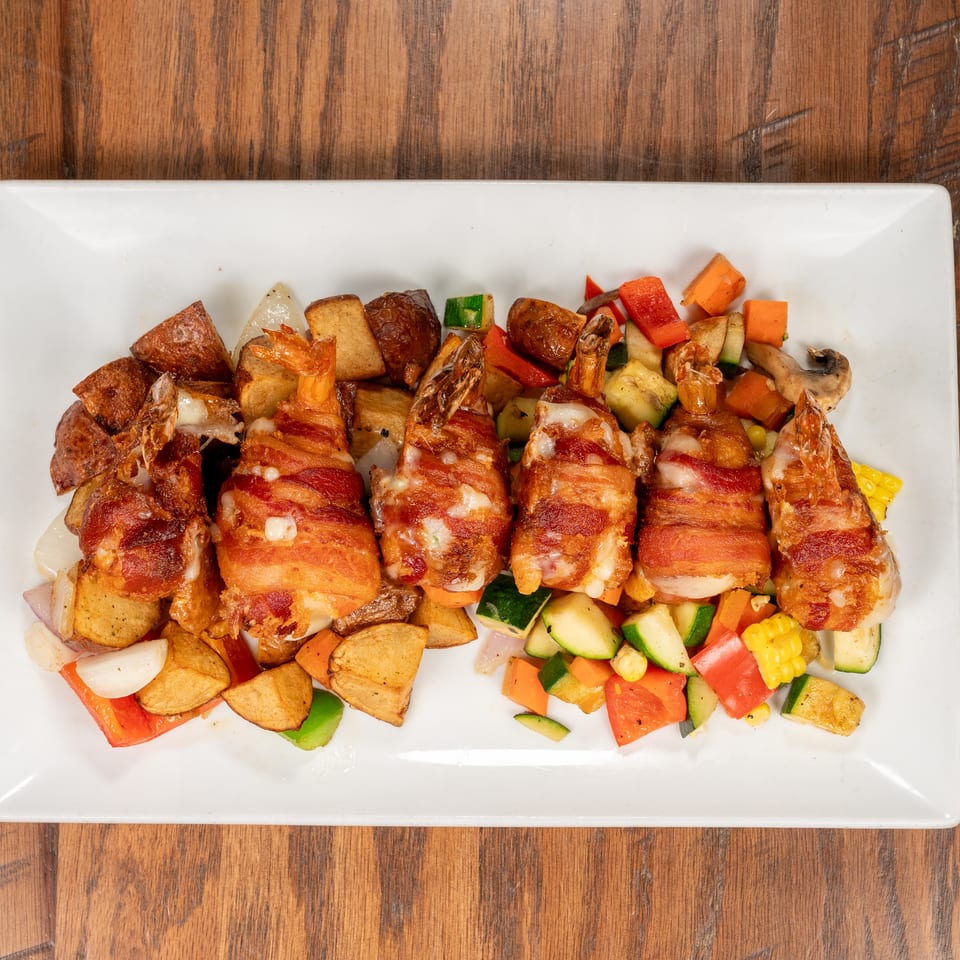 Bacon-wrapped shrimp with roasted potatoes and mixed vegetables on a white rectangular plate.