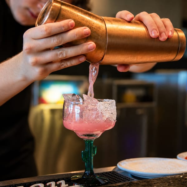 Bartender pouring a pink margarita from a copper shaker into a cactus-stemmed glass with a salted rim.