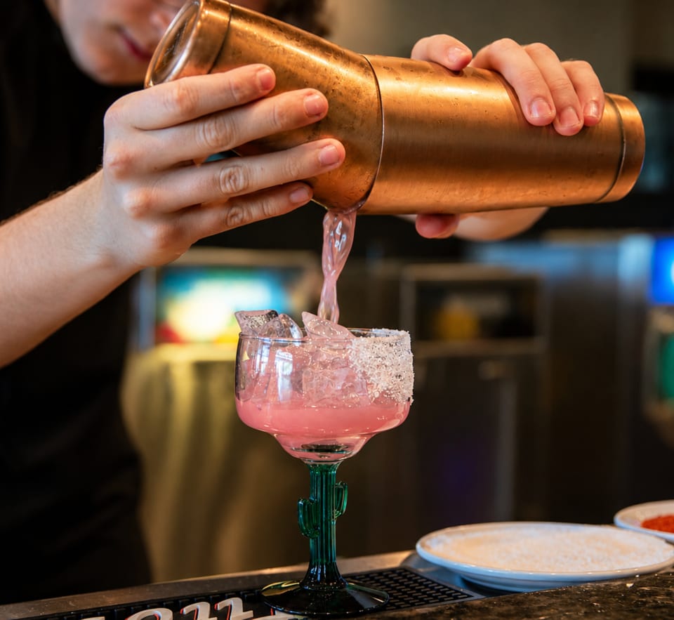 Bartender pouring a pink margarita from a copper shaker into a cactus-stemmed glass with a salted rim.