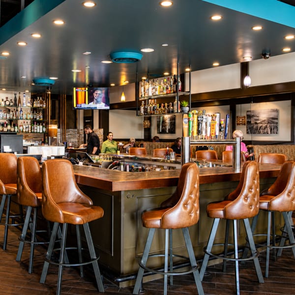A modern restaurant bar with brown leather stools, a dark wood counter, and bottles on shelves.