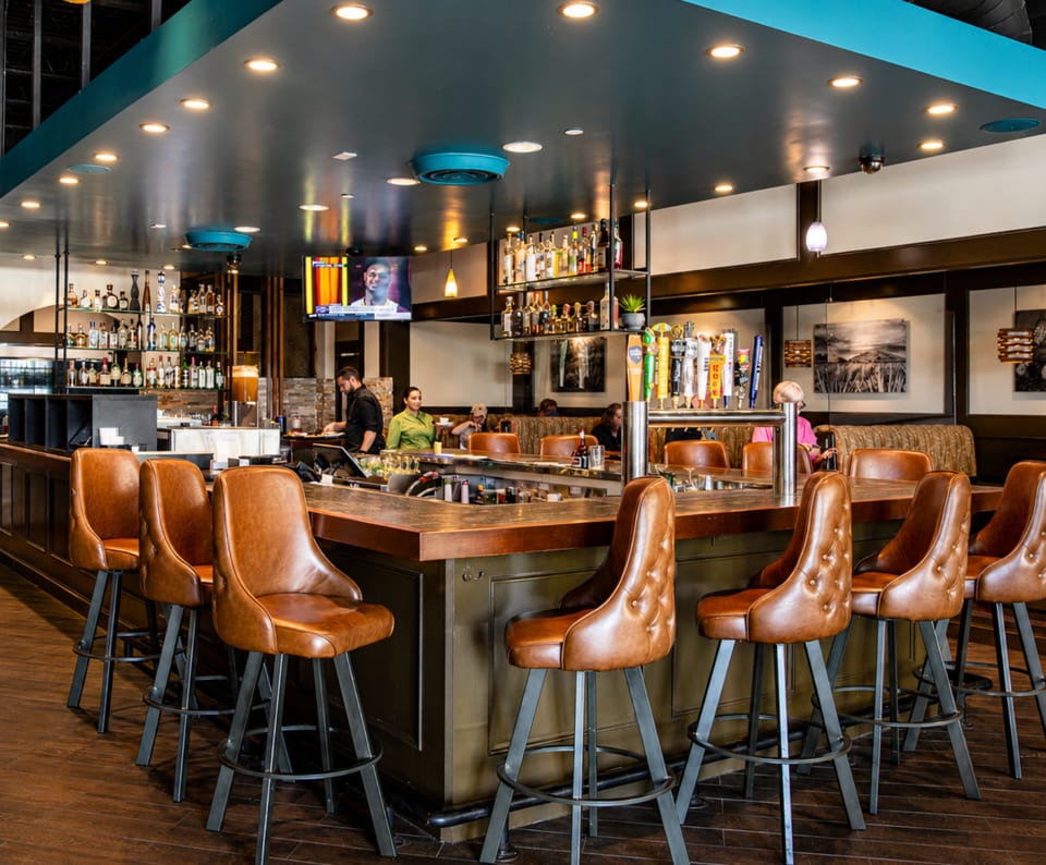 A modern restaurant bar with brown leather stools, a dark wood counter, and bottles on shelves.