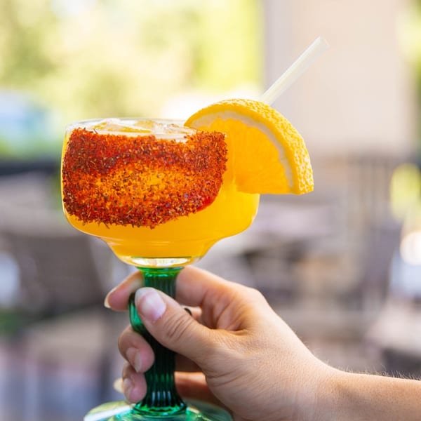 Hand holding a vibrant orange margarita in a green glass, rimmed with chili and garnished with an orange slice.