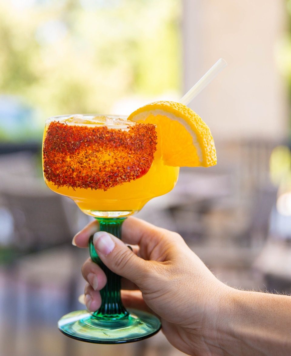 Hand holding a vibrant orange margarita in a green glass, rimmed with chili and garnished with an orange slice.