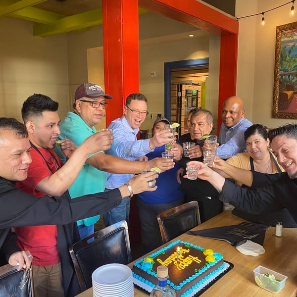 Restaurant staff and guests raise shot glasses in a toast around a "Happy Birthday" cake.