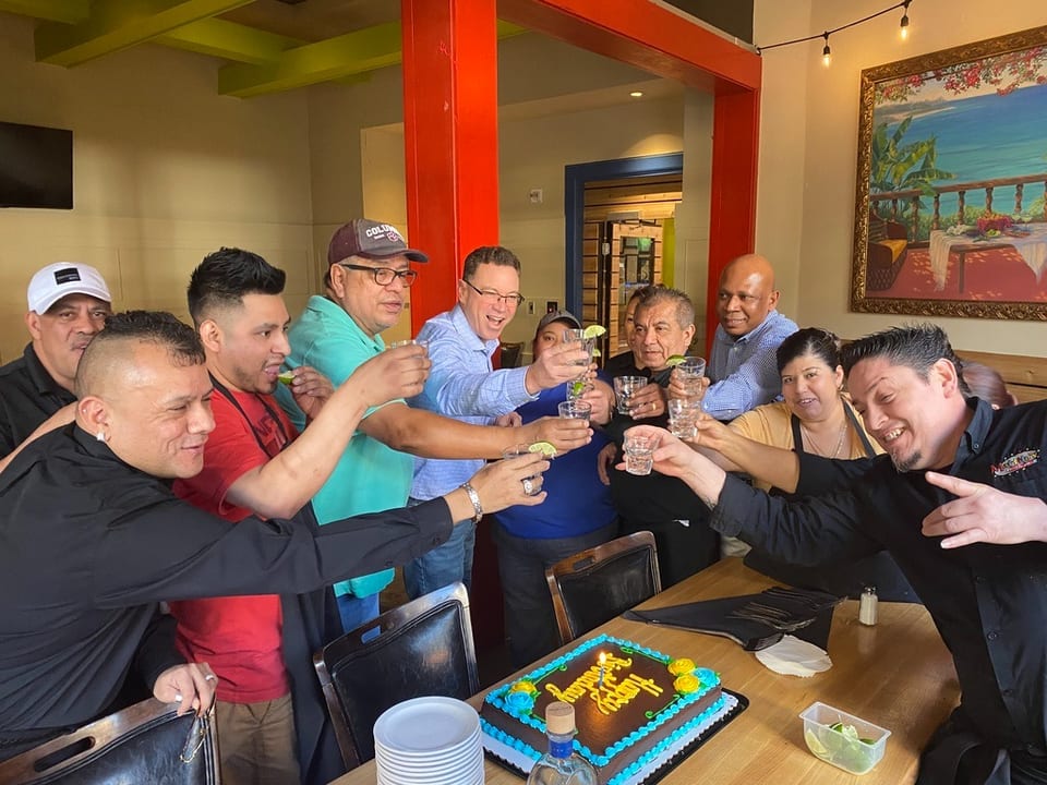 Restaurant staff and guests raise shot glasses in a toast around a "Happy Birthday" cake.
