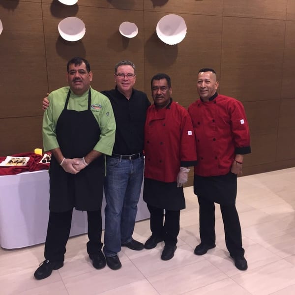 Four men, three in chef's coats, stand together in a restaurant dining area.