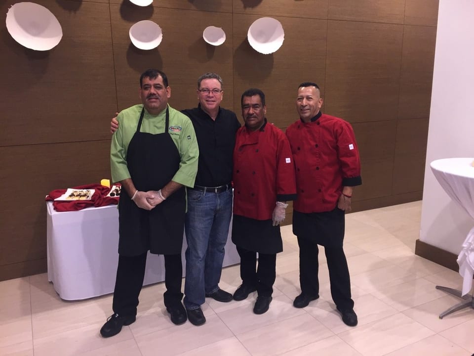 Four men, three in chef's coats, stand together in a restaurant dining area.