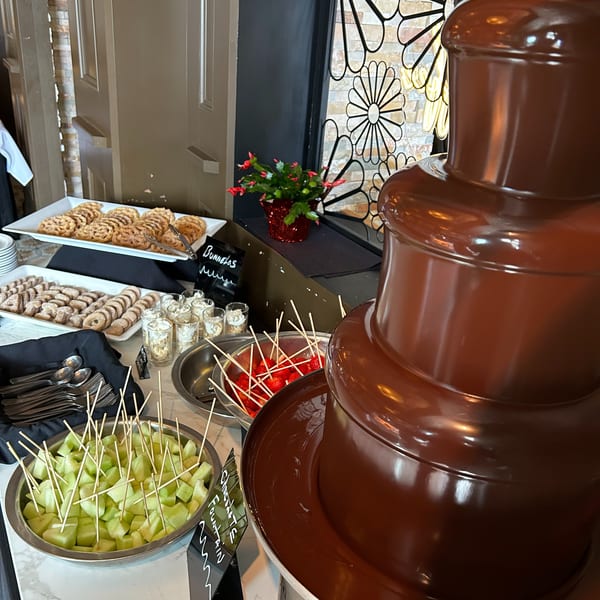 A chocolate fountain with various dipping items like melon, strawberries, cookies, and dessert cups.