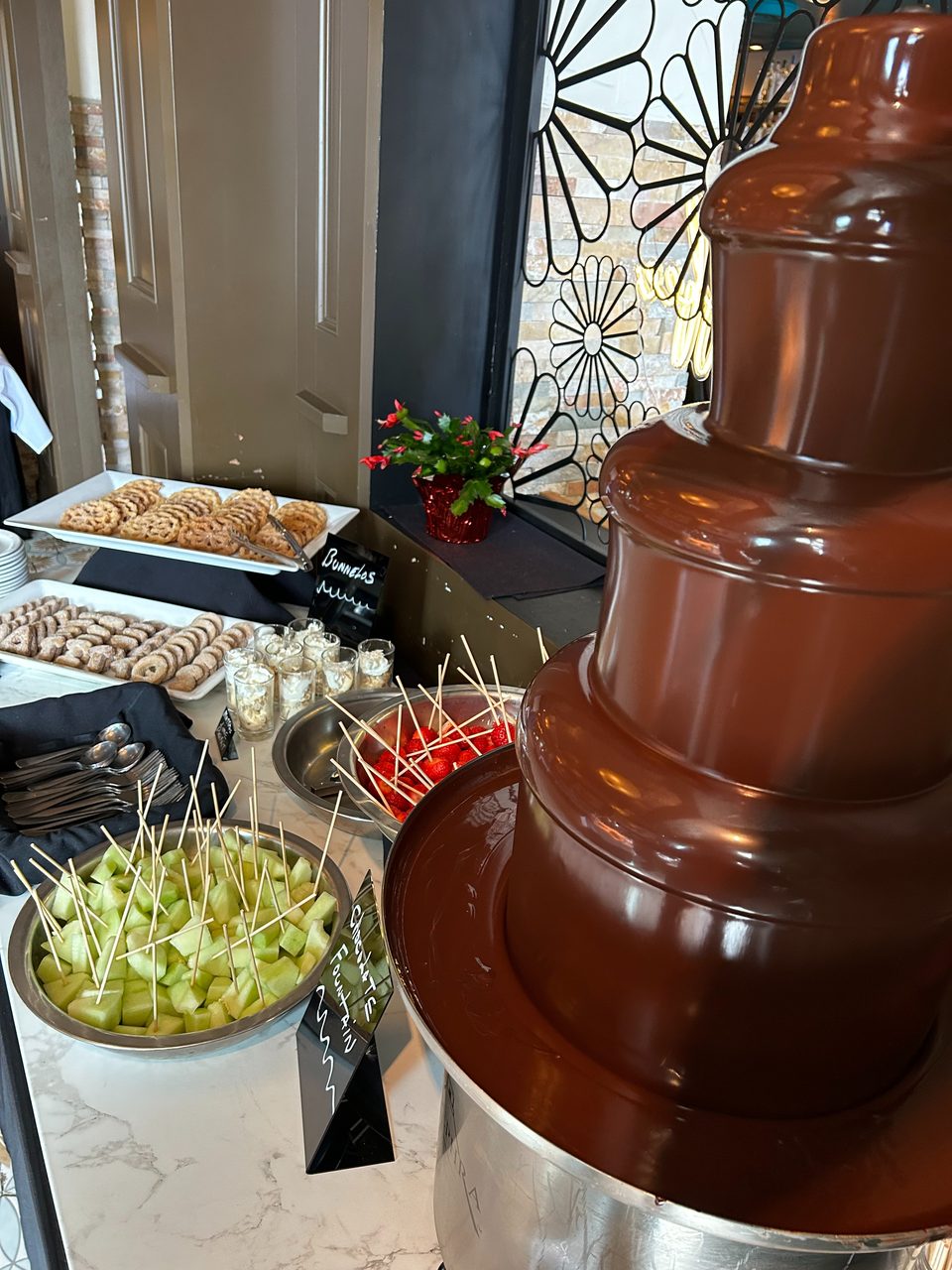 A chocolate fountain with various dipping items like melon, strawberries, cookies, and dessert cups.