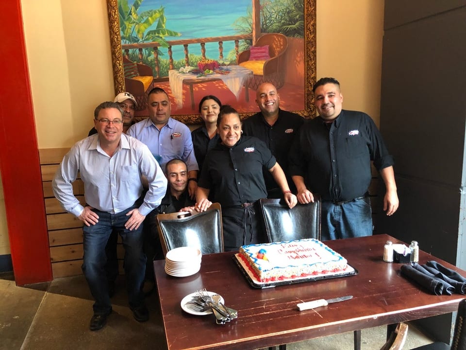 Restaurant staff gathered around a table with a birthday cake, smiling at the camera.