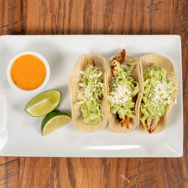 Three fish tacos with cabbage, cheese, and sauce, served with lime wedges and a side of orange sauce.