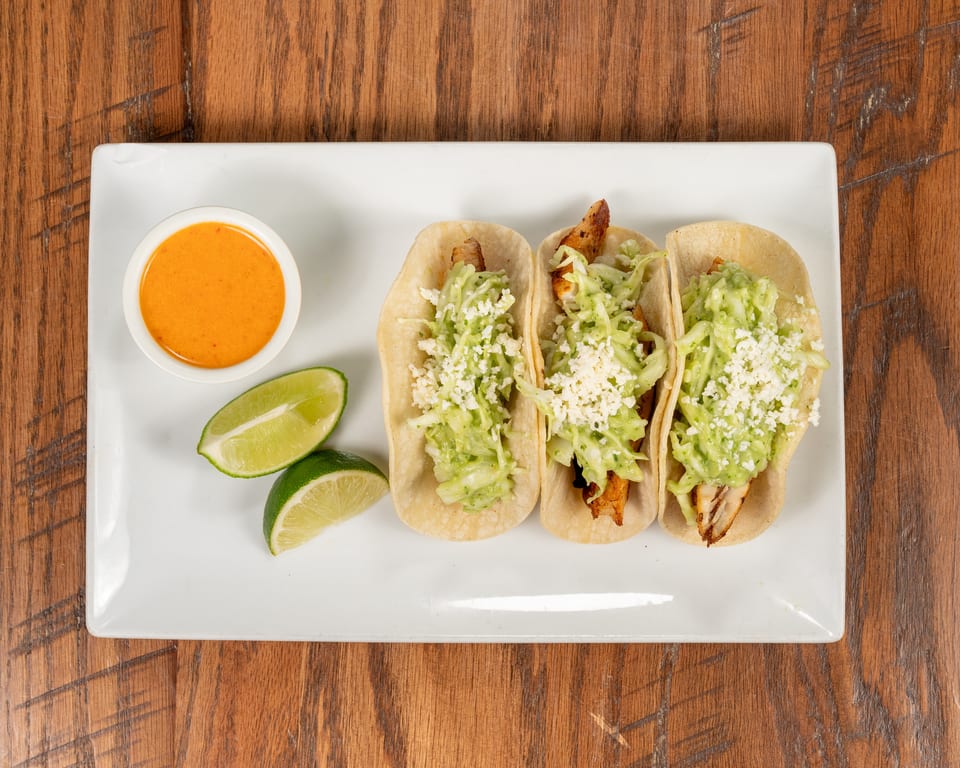 Three fish tacos with cabbage, cheese, and sauce, served with lime wedges and a side of orange sauce.