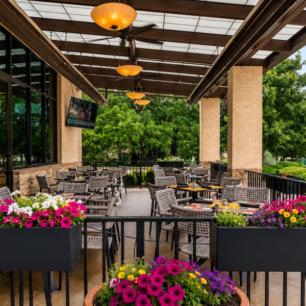 An outdoor patio with tables, chairs, and colorful flower planters, surrounded by lush green trees.