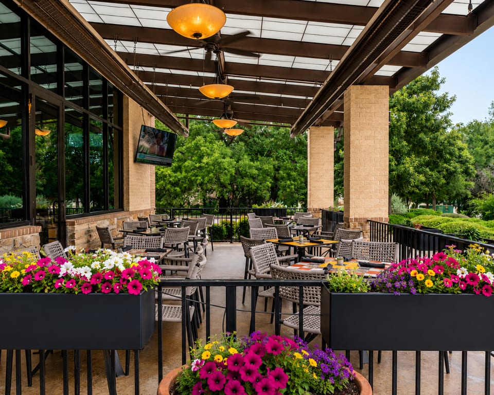 An outdoor patio with tables, chairs, and colorful flower planters, surrounded by lush green trees.
