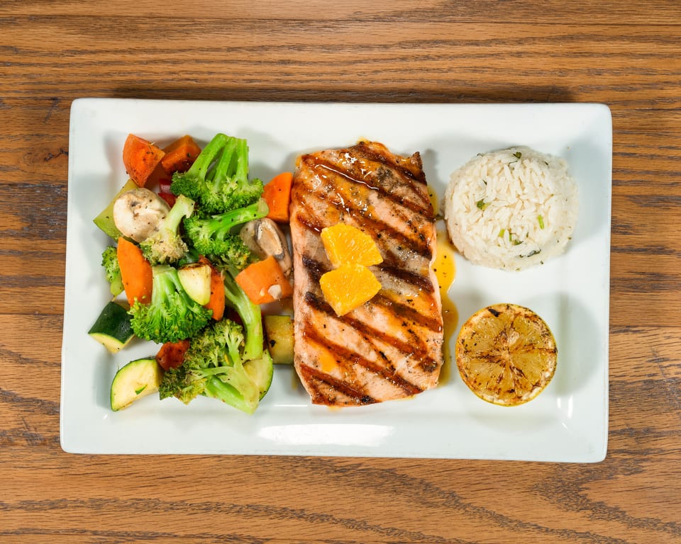 Grilled salmon with orange slices, roasted vegetables, rice, and a grilled lemon on a white plate.