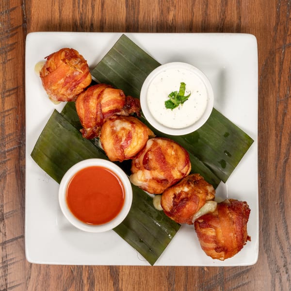 Bacon-wrapped appetizers with melted cheese, served with red and white dipping sauces on a white plate.