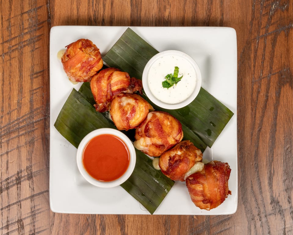 Bacon-wrapped appetizers with melted cheese, served with red and white dipping sauces on a white plate.
