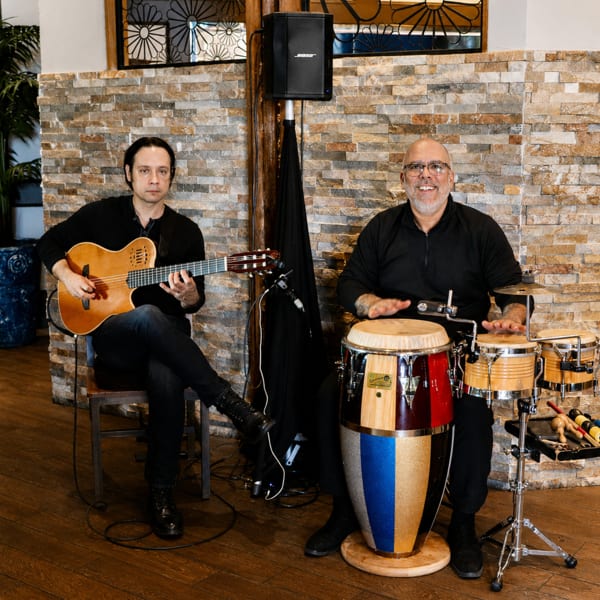Two musicians performing, one playing guitar and the other playing conga drums, in a restaurant.