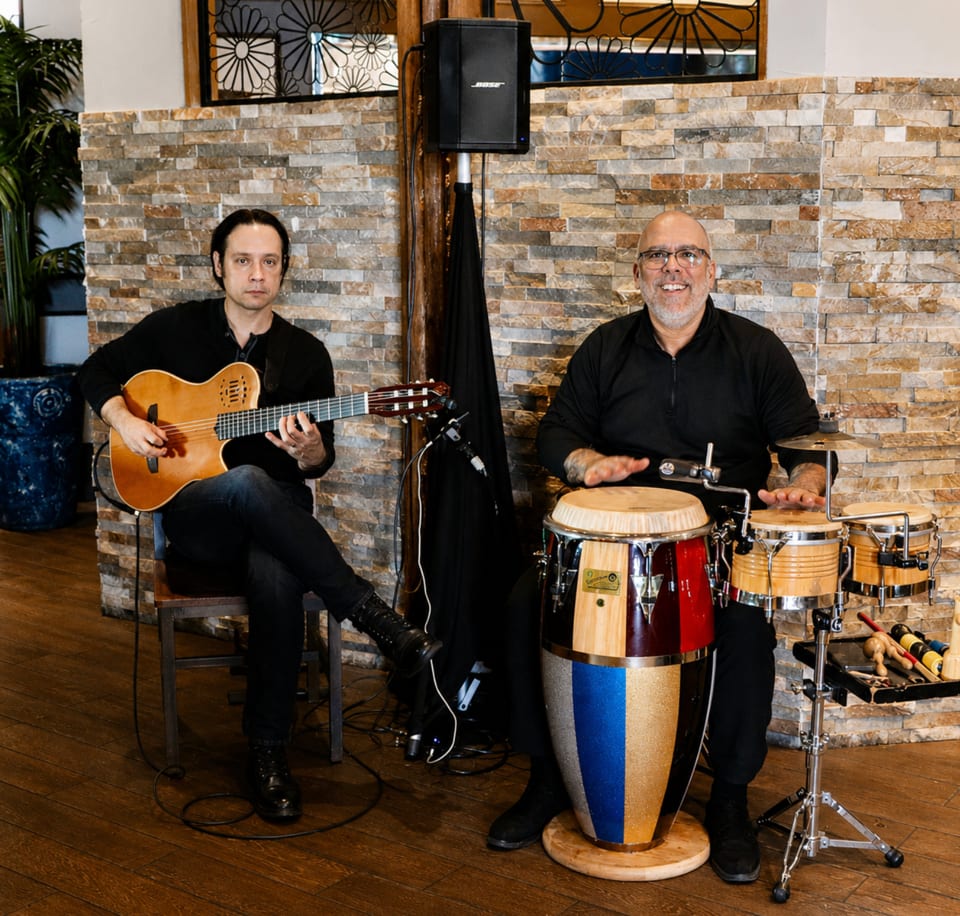 Two musicians performing, one playing guitar and the other playing conga drums, in a restaurant.