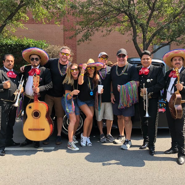 Mariachi band members and people in sombreros and Mardi Gras beads pose outdoors.