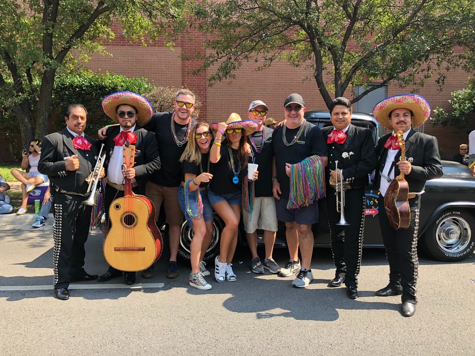 Mariachi band members and people in sombreros and Mardi Gras beads pose outdoors.