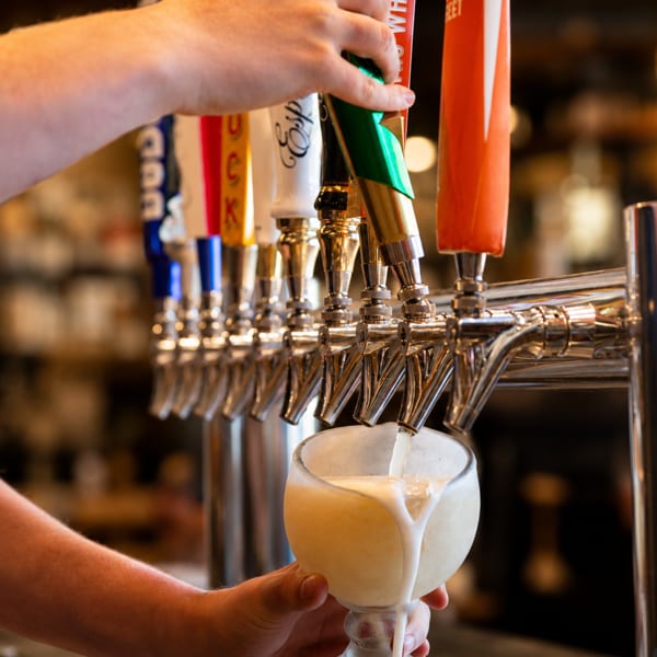 A bartender pours a frothy, light-colored drink from a tap into a glass.