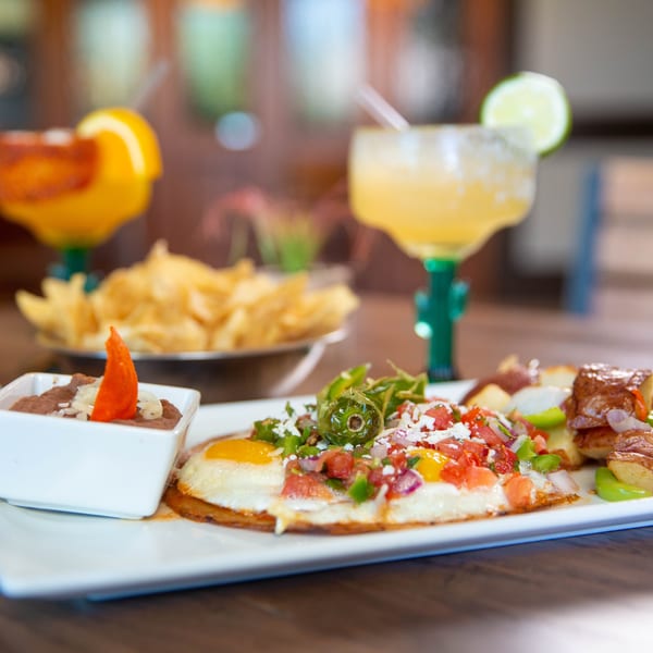 Huevos rancheros with refried beans, roasted potatoes, chips, and two margaritas on a wooden table.