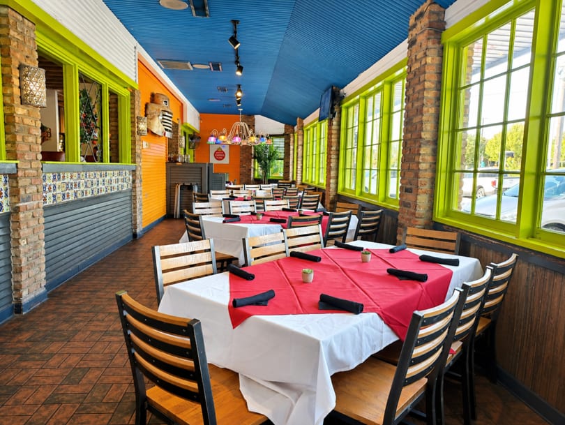 Vibrant restaurant interior with red and white tablecloths, wooden chairs, lime green window frames, and a blue ceiling.