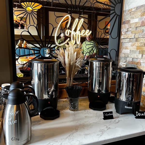 A coffee station with three large dispensers, a "Coffee" neon sign, and a half-and-half carafe.